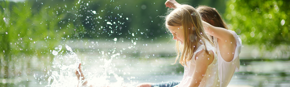 Two cute little girls sitting on a wooden platform by the river or lake dipping their feet in the water on warm summer day